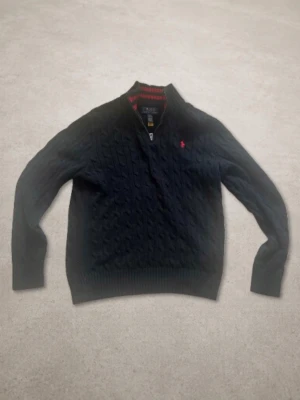 Mörkblå kabelstickad tröja Polo Ralph Lauren - Kabelstickad mörkblå tröja från Polo Ralph Lauren med halv dragkedja och röd broderad logga på bröstet. Tröjan har ribbade muddar och krage med röd detalj på insidan. OBS! Storleken är Junior Large (14-16) och motsvarar ungefär vuxen xs.
