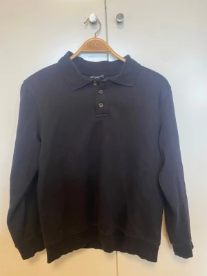 Svart långärmad polotopp med knappar - Klassisk svart långärmad polotopp med krage och tre knappar framtill. Tillverkad i mjukt material som känns skönt mot huden. Perfekt till jeans eller kjol för en clean och enkel look.