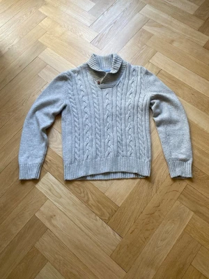 Ljusgrå kabelstickad tröja - Snygg ljusgrå kabelstickad tröja med polokrage och ribbade muddar. Tröjan har ett klassiskt flätmönster framtill och är perfekt för kyliga dagar. Materialet känns mjukt och skönt mot huden. Passar till både jeans och kjol.