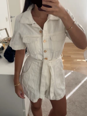 Vit kortärmad jumpsuit med bälte - Snygg vit jumpsuit med korta ärmar, knappar framtill i guld och matchande tygbälte i midjan. Två bröstfickor med lock och avslappnad krage. Perfekt till sommaren och gjord i ett lätt tyg som känns skönt mot huden. Aldrig använt.