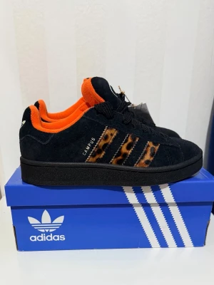 Adidas Campus svarta sneakers leopard - Svarta Adidas Campus sneakers med mockaöverdel, orange foder och leopardmönstrade ränder på sidorna. Klassisk Adidas-logga på plösen och svart sula. Snygg och unik design som sticker ut.