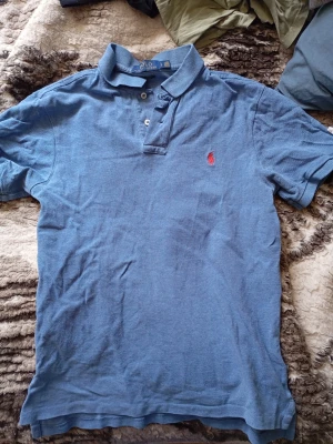 Blå Ralph Lauren pikétröja med krage - Snygg blå pikétröja med klassisk krage och knappar framtill. Tröjan har korta ärmar och en liten röd broderad logga på bröstet. Perfekt för en avslappnad och stilren look. Den är använd ganska mycket, men är fortfarande i bra skick. Inga hål eller fläckar. Pris kan diskuteras🙌. Mvh Rasmus.