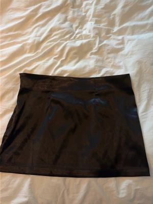 Svart kort satin-kjol - Snygg svart kortkjol i glansigt satinmaterial. Kjolen har en enkel och stilren design utan synliga detaljer, perfekt att matcha med olika toppar för en trendig look.