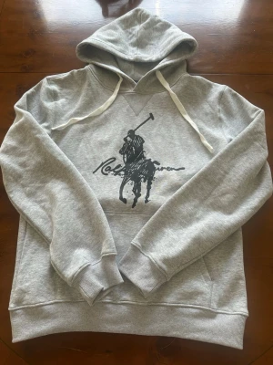 Grå Ralph lauren hoodie  - Snygg grå hoodie med stor svart Polo-inspirerad tryck på bröstet. Tröjan har huva med vita dragsnören och ribbade muddar vid ärmar och nederkant. Perfekt för en avslappnad och sportig stil. Tillverkad i mjukt sweatshirtmaterial.