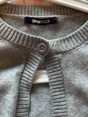Grå kofta från Gina Tricot - Klassisk grå kofta från Gina Tricot med rund halsringning och ribbade muddar. Koftan har en knapp framtill och är stickad i mjukt material. Perfekt att slänga över en t-shirt eller linne för en enkel och clean look.