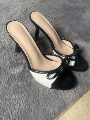 Svarta och vita pumps med rosett - Snygga pumps med öppen tå i svart och vitt. Skorna har en elegant rosett framtill och smala höga klackar. Ovandelen är i textil och syntet, vilket ger en stilren kontrast mellan färgerna. Perfekta för dig som vill ha en chic och trendig look.