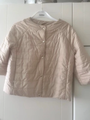Ljusbeige kort dunjacka - Snygg ljusbeige dunjacka med kort, boxig passform och trekvartsärmar. Jackan har rundad krage och stängs med silverfärgade tryckknappar framtill. Quiltad design och lätt vadderad för extra värme. Perfekt för kalla vinterdagar.