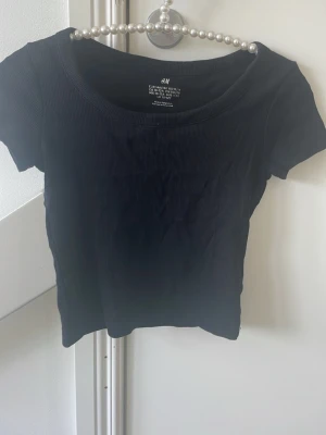 Svart ribbad topp från H&M - En svart ribbad topp från H&M med rund halsringning och korta ärmar. Toppen har en tight passform och är tillverkad i ett mjukt, stretchigt material som sitter snyggt på kroppen. Perfekt att matcha med jeans eller kjol.