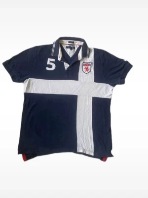 Marinblå Tommy Hilfiger pikétröja med vit kors - Snygg marinblå pikétröja med vit korsdetalj över bröstet och magen. Tröjan har klassisk krage med knappar, korta ärmar och en broderad sköld samt siffran 5 på bröstet. Perfekt för en sportig och avslappnad look.