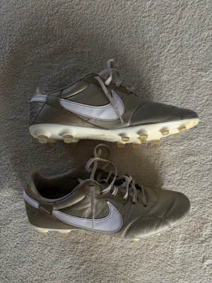 Beige Nike tiempos fotbollsskor med dobbar - Snygga beige fotbollsskor från Nike med vita detaljer och klassisk swoosh på sidan. Skorna har rundad tå, snörning och fasta dobbar i plast för spel på gräs. Ovandelen är i syntetmaterial och mesh för bra komfort och ventilation.