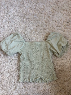 Ljusgrön topp med puffärmar Gina Tricot - Supersöt ljusgrön topp från Gina Tricot med smockad kropp och stora puffärmar. Toppen har en rak halsringning och volangkant nedtill. I storlek M. 