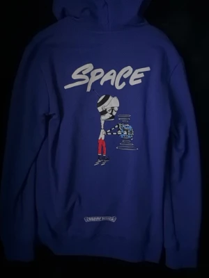 Blå chrome hearts hoodie med space-tryck - Cool blå hoodie med stor grafisk SPACE-print på ryggen och en tecknad figur i rymdtema. Tryck på båda ärmarna med eldflammor och tänder. Liten text på bröstet. Klassisk huva och känguruficka framtill. Perfekt för dig som gillar streetwear och unika prints.