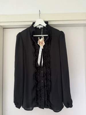 Svart blus med volangdetaljer - Elegant svart blus från Branting, perfekt för dig som uppskattar tidlös design. Denna oanvända topp i storlek M har fortfarande prislappen kvar och erbjuder en sofistikerad look med sina vackra volanger och klassiska snitt. Ett utmärkt val för att addera en touch av elegans till din garderob, vare sig det är till vardags eller fest.