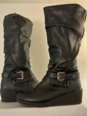  boots med kilklack - Säljer ett par svarta knähöga boots med kilklack och dekorativa spännen i metall. Skorna har dragkedja längs sidan och är gjorda i konstläder. Perfekta för dig som gillar en edgy stil och vill ha boots som sticker ut.
