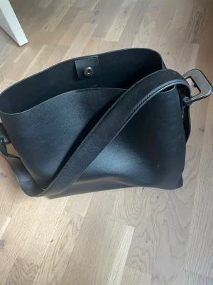 Svart axelväska i slätt konstläder - Stilren svart axelväska i slätt konstläder med bred axelrem och magnetknäppning. Väskan har en rymlig insida och ett innerfack med dragkedja för småprylar. Perfekt för dig som vill ha en enkel men snygg väska till vardagslooken.