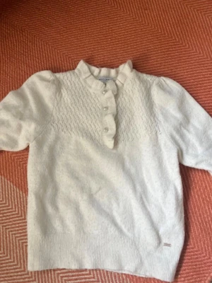 Vit stickad topp med puffärm - Söt vit stickad topp från Jacqueline de Yong med korta puffärmar och dekorativa knappar framtill. Tröjan har ett diskret mönster upptill och ribbade muddar vid ärmslut och nederkant. 