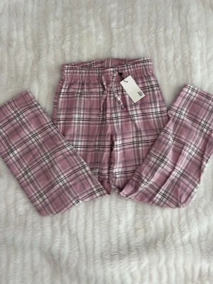Rosa rutiga pyjamasbyxor 140cl - Säljer ett par rosa pyjamasbyxor med vitt och svart rutmönster. Byxorna har resår i midjan och dragsko för bästa passform. Perfekta för mysiga kvällar hemma. Materialet känns mjukt och bekvämt mot huden.