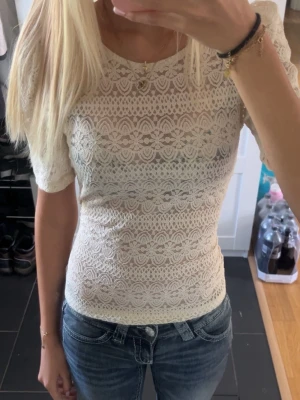 Vit spets tröja från Vila - Säljer en superfin vit spetsblus från Vila med korta puffärmar och rund hals. Blusen har ett detaljerat spetsmönster med blommor och knäppning bak i nacken. Perfekt att styla med jeans eller kjol för en trendig look. Säljer pga jag inte använder den❣️