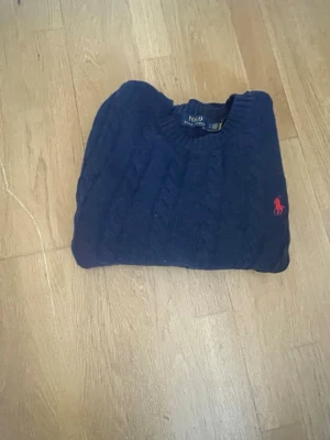 Mörkblå kabelstickad tröja Polo Ralph Lauren - Mörkblå kabelstickad tröja från Polo Ralph Lauren med rund halsringning och röd broderad logga på bröstet. Tröjan har långa ärmar och ribbade muddar vid ärmslut och nederkant. Klassisk och tidlös design i mjukt material. Storlek s.
