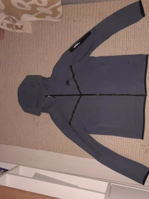 Grå nike tech  - Snygg grå vindjacka med hel dragkedja framtill och huva. Jackan har svarta detaljer vid ärmslut och fickor, samt en extra ficka med dragkedja på ena ärmen. Perfekt för blåsiga dagar och enkel att matcha.