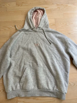 Grå hoodie med rosa rosett och fluffig huva - Mysig grå hoodie med en liten rosa rosett broderad på bröstet. Huvan är fodrad med ljusrosa fluffigt tyg och har dragsko. Klassisk känguruficka framtill och ribbade muddar vid ärmar och nederkant. Perfekt för chill dagar.