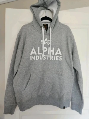 Hoodie Alpha Industries S - Grå hoodie Alpha Industries S Snygg grå hoodie från Alpha Industries med stor vit logotyp på bröstet och klassisk huva med snörning. Mjuk insida och ribbade muddar vid ärmar och nederkant. Perfekt för chill dagar och streetstyle.  Kan även hämtas I Partille  E1