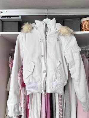 Vit pufferjacka med fuskpäls - Snygg vit pufferjacka med avtagbar huva och fuskpälsdetaljer. Jackan har stora knappar, dragkedja, ribbade muddar och fickor framtill samt en liten ficka på ärmen. Broderad logga på bröstet ger extra stilpoäng. Perfekt för kalla vinterdagar.