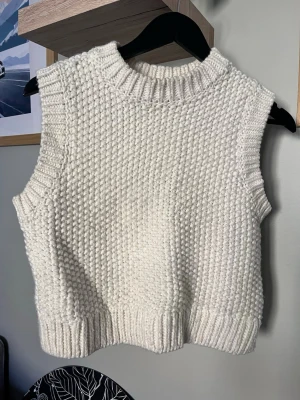 Vit stickad väst med grov struktur - Säljer en vit stickad väst med grov struktur och ribbade kanter vid hals, ärmhål och nederkant. Västen har en boxig passform och är perfekt att styla över en skjorta eller t-shirt för en trendig lager-på-lager-look.