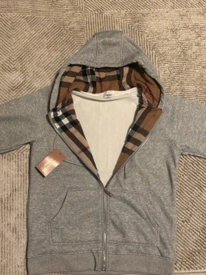 Grå zip up Burberry kofta  - Snygg grå hoodie med dragkedja och fickor framtill. Huvan har ett klassiskt rutigt foder i beige, svart och rött som ger en unik touch. Perfekt för dig som vill ha en stilren men ändå trendig look. Mjuk bomullskänsla och ribbade muddar.