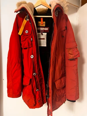 Röd parkas från Parajumpers - Säljer en röd Parajumpers parkas med huva och fuskpäls, flera fickor och detaljer som stora knappar och dragkedjor. Jackan har en gul rem vid halsen och är fodrad för extra värme. Perfekt för kalla dagar och riktigt snygg streetstil.