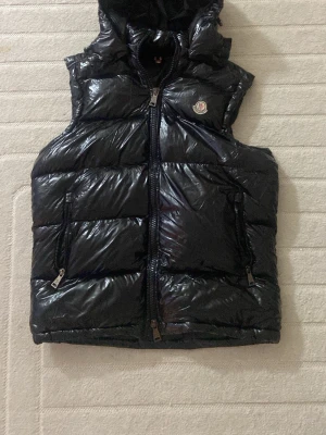 Svart glansig moncler  dunväst med huva - Snygg svart dunväst från Moncler med glansigt material och avtagbar huva. Västen har två dragkedjeförsedda fickor framtill och en klassisk Moncler-logga på bröstet. Perfekt för lager-på-lager och streetstyle.