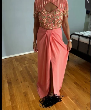 Lång rosa klänning med öppen rygg - Elegant långklänning i rosa med blommiga broderier och paljetter upptill. Klänningen har korta ärmar, öppen rygg och en slits framtill. Perfekt för bal eller festliga tillfällen.