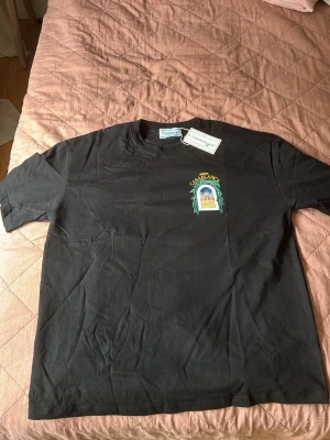Svart t-shirt med Casablanca-tryck - Cool svart t-shirt med färgglatt tryck på bröstet där det står 'Avenida Casablanca' och ett motiv av en byggnad i fönster med gröna blad runt. T-shirten har rund hals och korta ärmar, perfekt för en avslappnad streetwear-look.