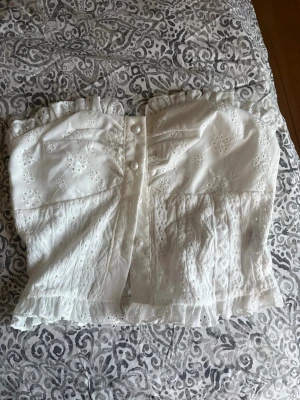 Vit spetsig axelbandslös korsettopp - Supersöt vit axelbandslös korsettopp med spetsdetaljer och broderade blommor. Toppen har volangkant upptill och nedtill samt knappar framtill för extra stil. Perfekt för dig som vill ha en romantisk och trendig look i sommar.