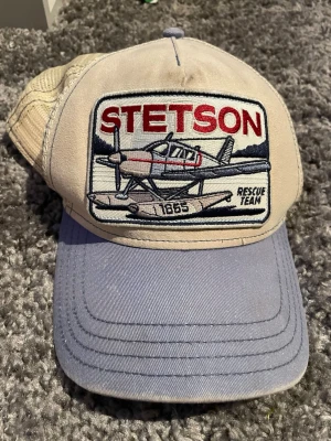 Beige och blå Stetson keps - Säljer en beige och blå keps från Stetson med broderad patch framtill som visar ett sjöflygplan och texten 'Rescue Team'. Kepsen har mesh baktill och justerbar snapback i blå plast. Perfekt för dig som gillar retrostil och detaljerade broderier.