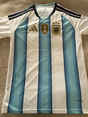 Argentina matchtröja Adidas blåvit - Säljer en officiell Argentina matchtröja från Adidas med klassiska blå och vita ränder, guldiga detaljer och FIFA-märke på bröstet. Tröjan har korta ärmar, svart axelparti med tre ränder och är tillverkad i lätt, ventilerande material.
