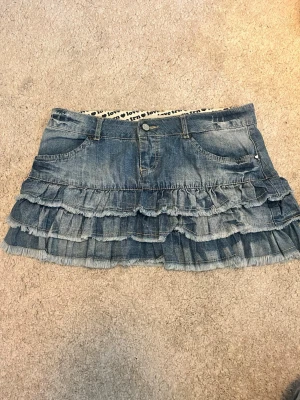 Blå jeanskjol med volanger - Säljer en kort jeanskjol i blå denim med flera lager volanger och fransade kanter. Kjolen har klassiska fickor fram och bak samt knapp och dragkedja i midjan. Perfekt för dig som gillar en cool och avslappnad stil.💞 passar som S