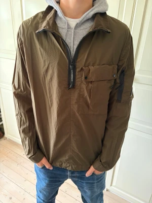 Brun overshirt från C.P. Company - Snygg brun overshirt från C.P. Company med dragkedja framtill, bröstficka med knapp och en extra ficka med dragkedja på ärmen. Tillverkad i lätt, krispigt material och har den klassiska C.P Company-loggan på ärmen. Jackan är använd max 2-3 gånger.Jackan är i storlek L. Hör av er vid frågor och priset är inte hugget i sten!