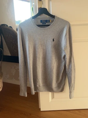 Grå tröja från Polo Ralph Lauren - Klassisk grå tröja från Polo Ralph Lauren med rund halsringning och den ikoniska broderade loggan på bröstet. Tillverkad i mjukt material och har ribbade muddar vid ärmslut och nederkant. Perfekt för en clean och stilren look.