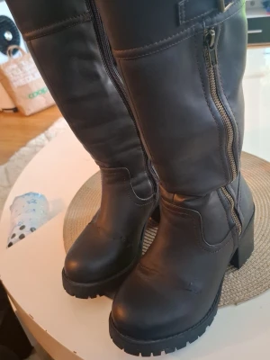 Svarta höga stövlar med grov sula - Snygga svarta stövlar i skinnimitation med grov klack och dragkedja på sidan. Stövlarna har en hög modell som går upp till knäet och en markerad sula för extra grepp. Perfekta till höstens och vinterns outfits.