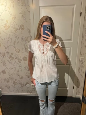 Vit blus med spetsdetaljer - Söt vit blus med korta ärmar och v-ringning dekorerad med spets och knytning framtill. Blusen har en luftig passform och tunnt, lätt material som ger en romantisk känsla. Perfekt att matcha med jeans för en avslappnad men ändå stilsäker look. Använd fåtal gånger och är i bra skick. 