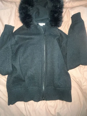 Svart stickad hoodie från Gina Tricot - Mysig svart stickad hoodie från Gina Tricot med dragkedja framtill och huva med fluffig fuskpälsdetalj. Tröjan har långa ärmar och ribbade muddar vid ärmslut och nederkant. Perfekt för kyliga dagar!