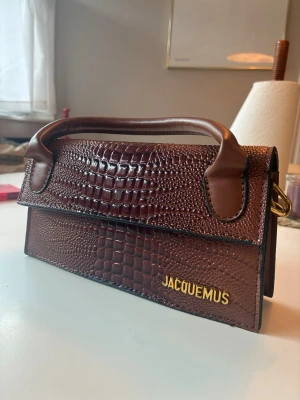 Jacquemus - Säljer en brun handväska i croco-präglat skinn med guldfärgad logga framtill. Väskan har ett stilrent handtag och en rektangulär form som ger en trendig look. Perfekt accessoar för dig som vill sticka ut med en lyxig touch.