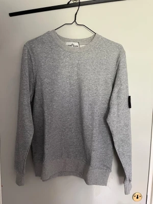 Grå tröja Stone Island - Klassisk grå sweatshirt från Stone Island med rund halsringning och ribbade muddar. Tröjan har den ikoniska svarta logotypen på vänster ärm. Perfekt för en avslappnad och stilren look.