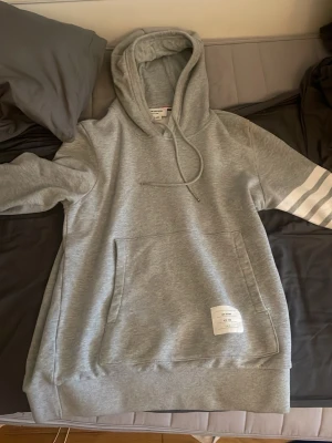 Thom Browne hoodie Size 2 - Snygg grå hoodie från Thom Browne med klassiska vita ränder på ena ärmen och stor magficka. Hoodien har dragsko i huvan och en patch med logga framtill. Perfekt för en chill och stilren look.