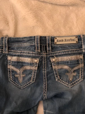 Rock revival lågmidjade jeans  - Blåa lågmidjade rock revival bootcut jeans med vita sömmar och så snygga glitter fickor! Stl 26 som motsvarar xs/s skulle jag säga. Avklippta nån ynka centimer på längden, skriv för exakta mått osv! 💘