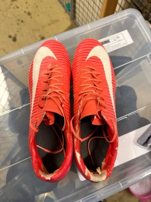 Röda Nike fotbollsskor med dobbar - Säljer ett par röda Nike fotbollsskor med vita detaljer och klassisk swoosh på sidorna. Skorna har snörning och dobbar för bra grepp på planen. Tillverkade i syntetmaterial med ribbad ovandel för bättre bollkontroll. Perfekta för dig som vill sticka ut på fotbollsplanen.
