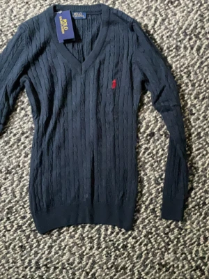 Mörkblå kabelstickad tröja Polo Ralph Lauren - Mörkblå kabelstickad tröja från Polo Ralph Lauren med V-ringning och lång ärm. Tröjan har det klassiska röda broderade logot på bröstet och ribbade muddar vid ärmslut och nederkant. Perfekt för en stilren och tidlös look.