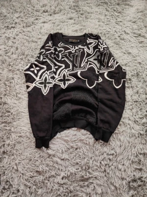 Svart sweatshirt - Cool svart sweatshirt med stora vita loggor och monogrammönster över hela tröjan. Rund halsringning och ribbade muddar vid ärmslut och nederkant. Perfekt för dig som vill sticka ut med en exklusiv streetwear-look. Säljer pågrund av att tröjan inte passar mig. Ställ frågor och priset är diskuterbart!