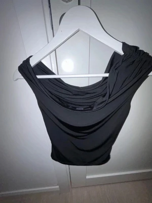 Svart offshoulder topp med drapering - Säljer en svart offshoulder topp med snygg drapering framtill. Toppen är ärmlös och har en croppad passform som ger en trendig look.  man kan ha den på fest eller bara till vardags. Aldrig använd ändats testad.